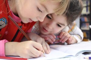Fotografische Darstellung zweier Kinder im Grundschulalter. Die Kinder sitzen nebeneinander an einem Tisch. Ein Kind malt auf einem Notizblock, das andere Kind schaut dabei zu.