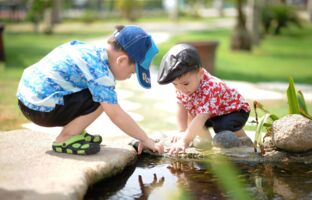 Fotografische Darstellung zweier Jungen im Kleinkindalter. Die Jungen spielen in gehockter Position an einem Wasserlauf.